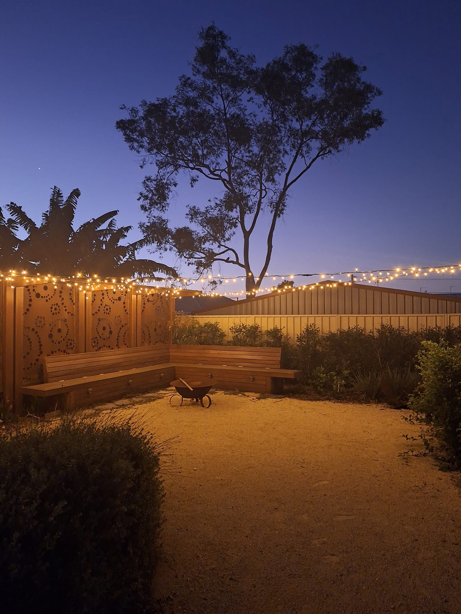 Backyard courtyard at dusk lit with warm festoon string lights and uplit decorative screens